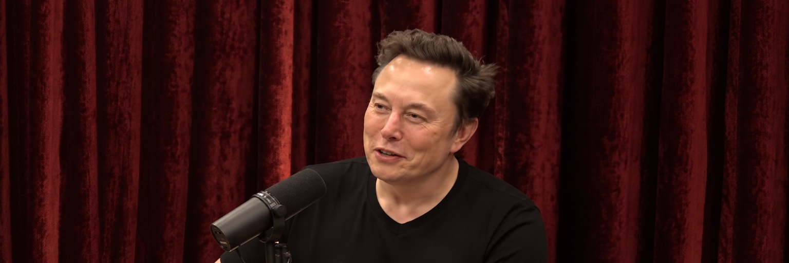 Elon Musk speaking on a podcast in front of a red curtain